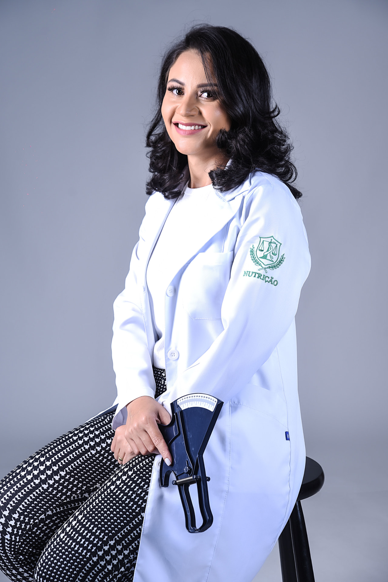 Daniela Ramos sorrindo, de jaleco branco, sentada em estúdio